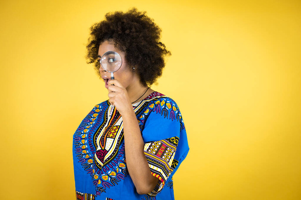 Femme afro-américaine portant des vêtements africains sur fond jaune surpris regardant à travers une loupe - Photo, image