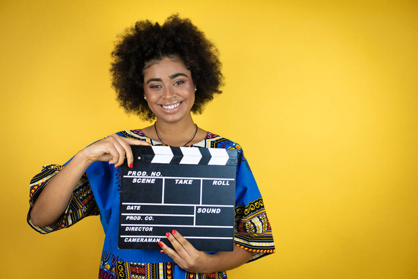 Африканская американка в африканской одежде на жёлтом фоне, держа планшет, очень счастлива веселиться. - Фото, изображение