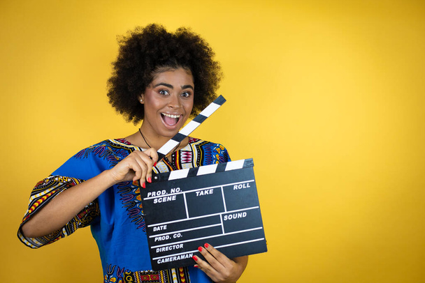 Sarı arka plan üzerinde Afrikalı giysiler giyen Afro-Amerikalı bir kadın elinde klaket tutarken çok mutlu ve eğleniyor. - Fotoğraf, Görsel