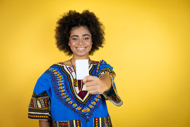 Αφροαμερικανή γυναίκα που φοράει αφρικανικά ρούχα πάνω από κίτρινο φόντο χαμογελώντας και κρατώντας λευκή κάρτα - Φωτογραφία, εικόνα