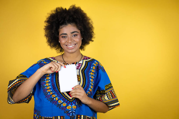 Afroamerykanka ubrana w afrykańską odzież na żółtym tle uśmiechnięta i z pustym notatnikiem w ręku - Zdjęcie, obraz