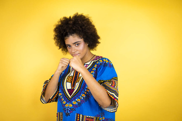 Afro-Amerikaanse vrouw draagt Afrikaanse kleding over gele achtergrond ponsen vuist om te vechten, agressieve en boze aanval, bedreiging en geweld - Foto, afbeelding
