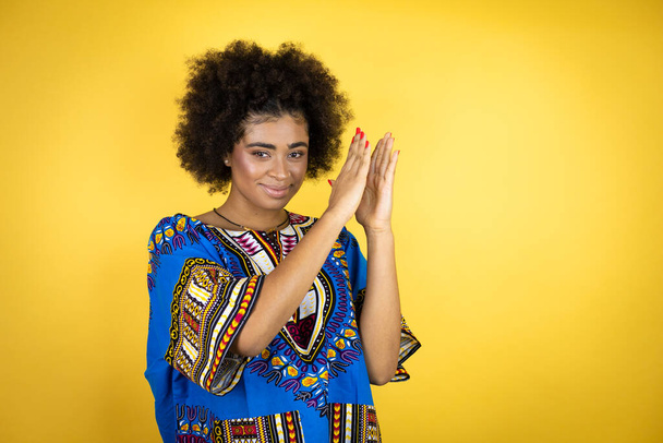 黄色の背景の上にアフリカの服を着てアフリカ系アメリカ人の女性が拍手と幸せと喜び、一緒に誇りに思って手を笑って - 写真・画像