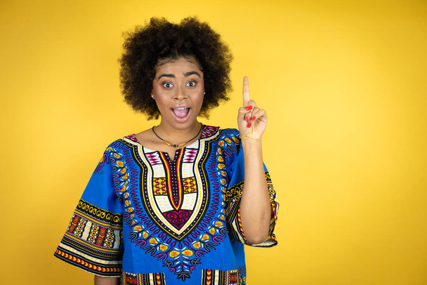 黄色の背景にアフリカの服を着たアフリカ系アメリカ人女性は、成功したアイデアで指を指しています。興奮し、幸せ。第一番. - 写真・画像