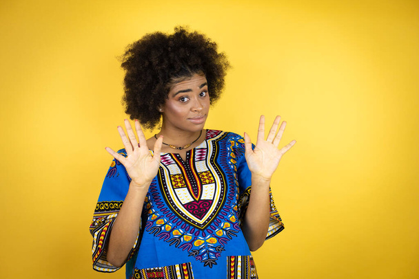 Mujer afroamericana vistiendo ropa africana sobre fondo amarillo disgustada con su mano dentro de su boca - Foto, Imagen