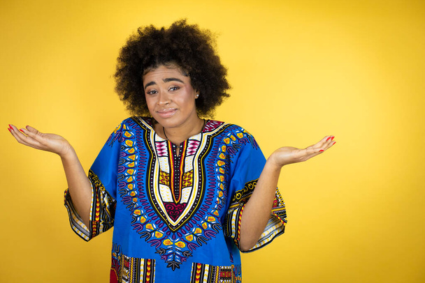 Afrikkalainen amerikkalainen nainen yllään afrikkalainen vaatteet yli keltainen tausta tietämätön ja sekava ilme kädet ja kädet ylhäällä - Valokuva, kuva