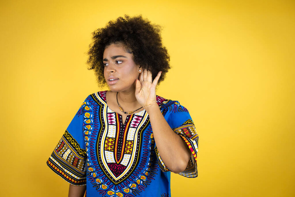 Afroamerikanerin in afrikanischer Kleidung vor gelbem Hintergrund überrascht mit Hand über Ohr, wenn sie Gerüchten oder Klatsch lauscht - Foto, Bild