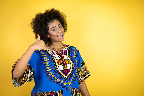 Afrikkalainen amerikkalainen nainen yllään afrikkalainen vaatteet keltainen tausta hymyillen tekee puhelimen ele käsin ja sormin kuin puhuminen puhelimessa - Valokuva, kuva