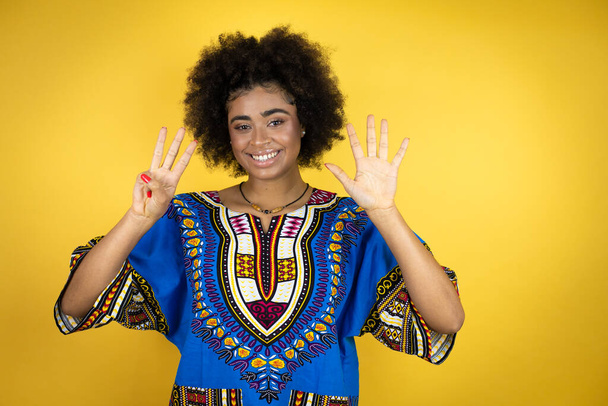 Afroamerikanerin in afrikanischer Kleidung vor gelbem Hintergrund zeigt mit Finger Nummer acht nach oben und lächelt dabei selbstbewusst und glücklich - Foto, Bild