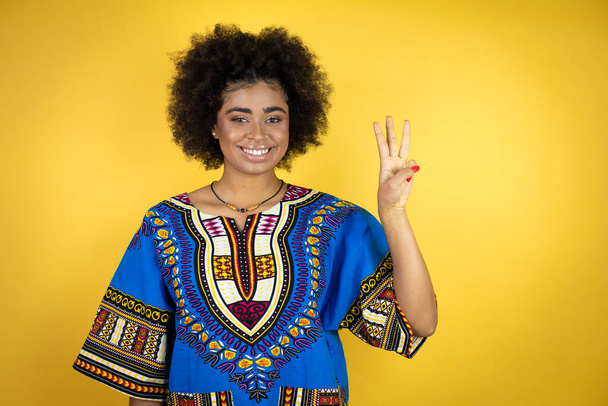 Afroamerikanerin in afrikanischer Kleidung vor gelbem Hintergrund zeigt mit Finger Nummer drei nach oben und lächelt dabei selbstbewusst und glücklich - Foto, Bild
