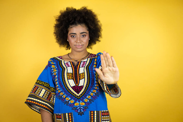 Mujer afroamericana vistiendo ropa africana sobre fondo amarillo serio y haciendo dejar de cantar con la palma de la mano. - Foto, Imagen