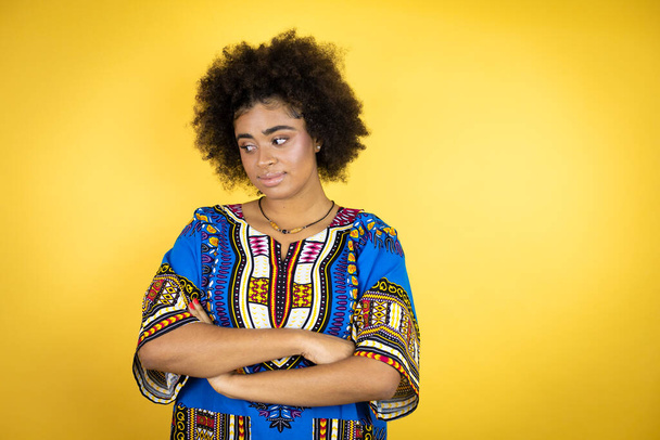Afroamerykanka ubrana w afrykańską odzież na żółtym tle, myśląca zmęczona i znudzona skrzyżowanymi ramionami. - Zdjęcie, obraz