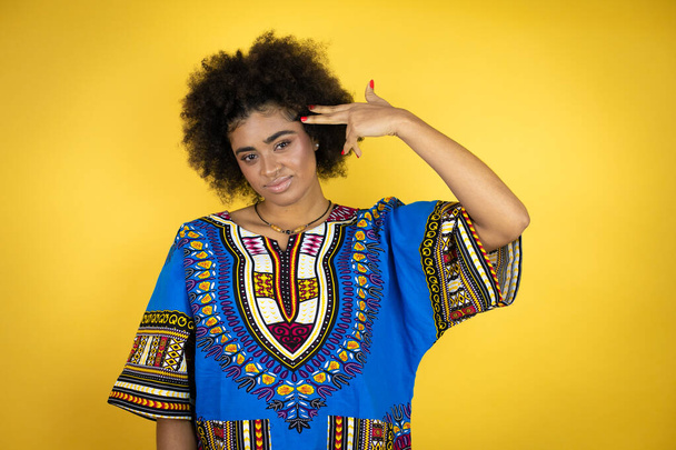 Mujer afroamericana vistiendo ropa africana sobre fondo amarillo disparándose y matándose apuntando de la mano y dedos a la cabeza como arma, gesto suicida. - Foto, Imagen