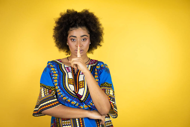 Afroamerykanka w afrykańskim ubraniu na żółtym tle prosi o ciszę z palcem na ustach. Cisza i tajna koncepcja. - Zdjęcie, obraz