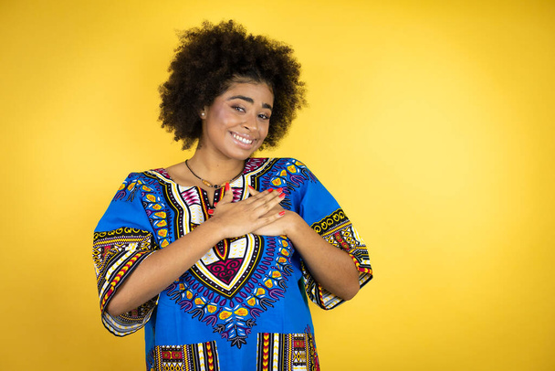 Mujer afroamericana vistiendo ropa africana sobre fondo amarillo sonriendo con las manos en el pecho y gesto agradecido en la cara. - Foto, Imagen