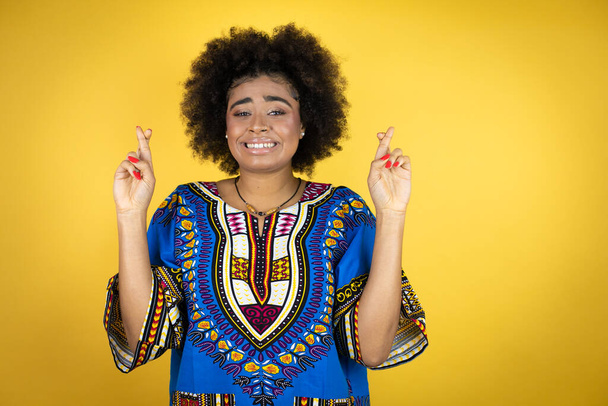 Afroamerikanerin in afrikanischer Kleidung vor gelbem Hintergrund gestikuliert mit erhobenem Zeigefinger lächelnd vor Hoffnung und schaut Seite an Seite - Foto, Bild