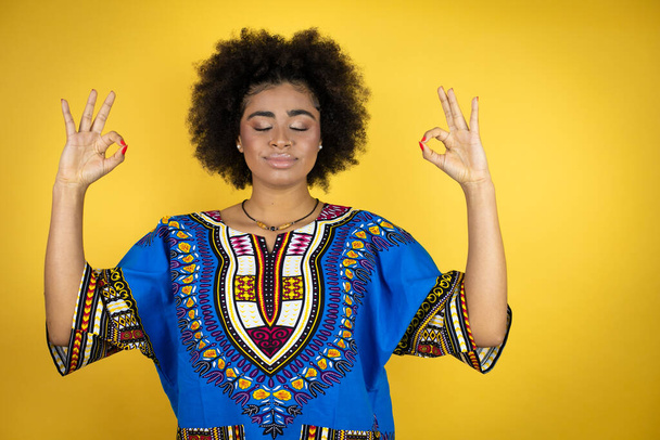 アフリカ系アメリカ人女性は黄色の背景の上にアフリカの服を着てリラックスし、目を閉じて笑顔指で瞑想ジェスチャーを行う - 写真・画像