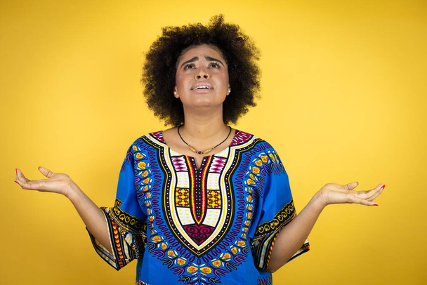 Afrikai nő afrikai ruhát visel sárga háttér tudatlan és zavaros kifejezés a karok és a kezek emelt - Fotó, kép