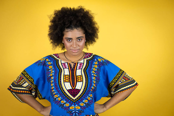 Afrika kökenli Amerikalı bir kadın sarı arka plan üzerine Afrikalı giysiler giyiyor. Kuşkucu ve gergin. Yüzünde, belinde kollarıyla onaylamayan bir ifade var. - Fotoğraf, Görsel