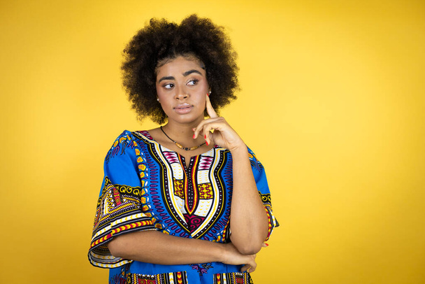 Mujer afroamericana vistiendo ropa africana sobre fondo amarillo cara seria pensando en la pregunta con la mano en la barbilla, pensativo acerca de la idea confusa - Foto, Imagen