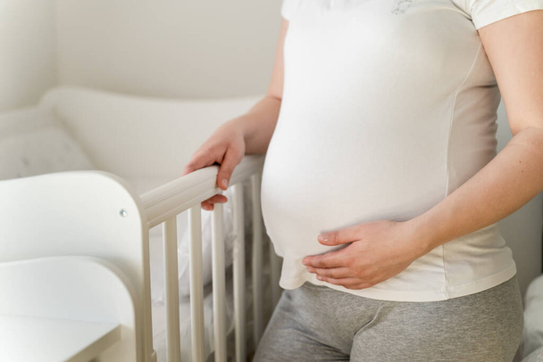 Κοντινό πλάνο στο μέσο της άγνωστης καυκασίας έγκυος γυναίκα στέκεται δίπλα στην κούνια σε φωτεινό δωμάτιο στο σπίτι κρατώντας την κοιλιά της - έννοια της μητρότητας και της εγκυμοσύνης - Φωτογραφία, εικόνα