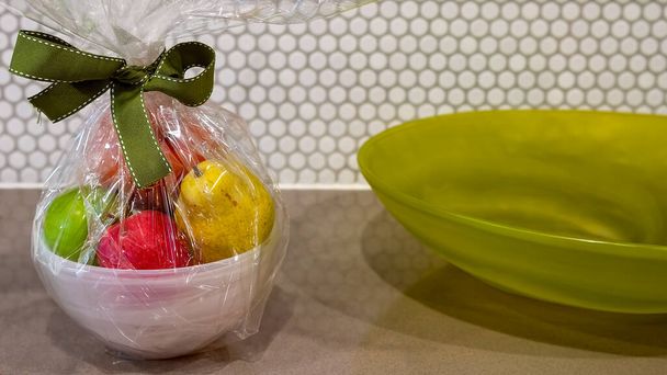 セロハンに包まれ、弓で結ばれたボウルに果物,装飾的な緑のガラス皿の横に. - 写真・画像