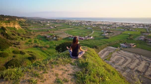 Mujer practica yoga y medita en la cima de la montaña con hermosas vistas de la ciudad y el océano al atardecer. Mujer sentada en pose fácil o sukhasana con mudra. Relajación, armonía con la naturaleza - Metraje, vídeo