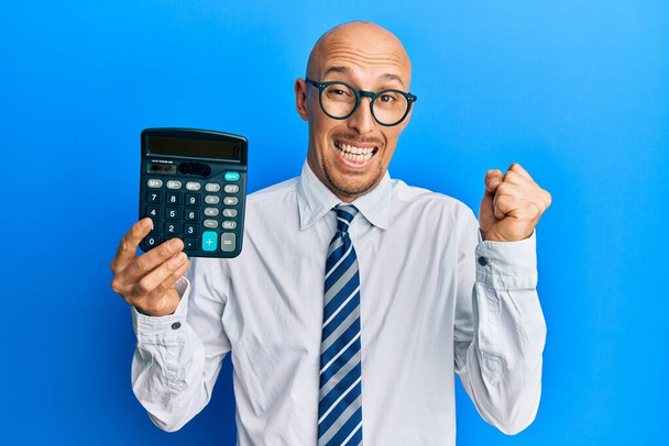 Bald business man with beard showing calculator device screaming proud, celebrating victory and success very excited with raised arm  - Photo, Image