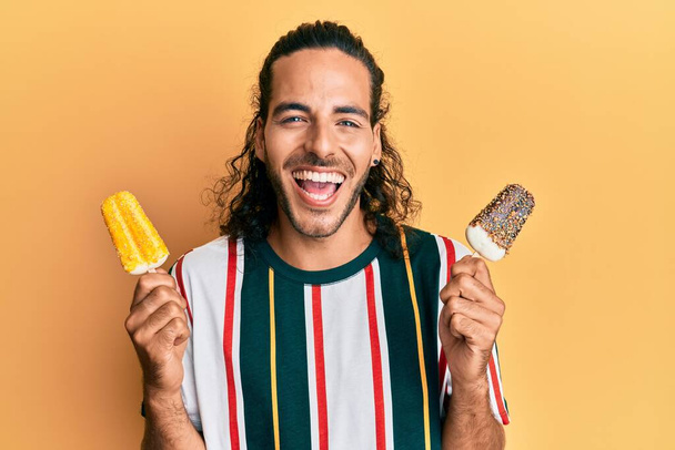 Молодий красивий чоловік з довгим волоссям їсть два морозива посміхається і сміється важко вголос, тому що смішний божевільний жарт
.  - Фото, зображення