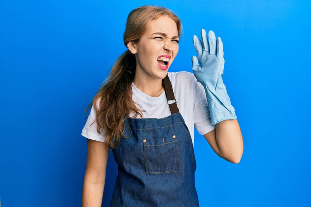 Beautiful blonde caucasian woman wearing cleaner apron and gloves shouting and screaming loud to side with hand on mouth. communication concept.  - Photo, Image