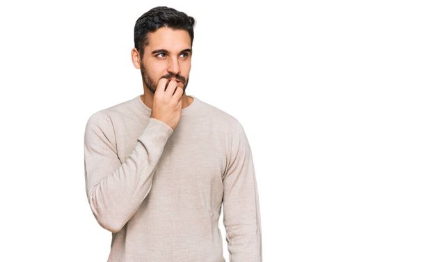 Jonge Spaanse man draagt casual kleding die er gestrest en nerveus uitziet met handen op de mond bijtende nagels. angstprobleem.  - Foto, afbeelding