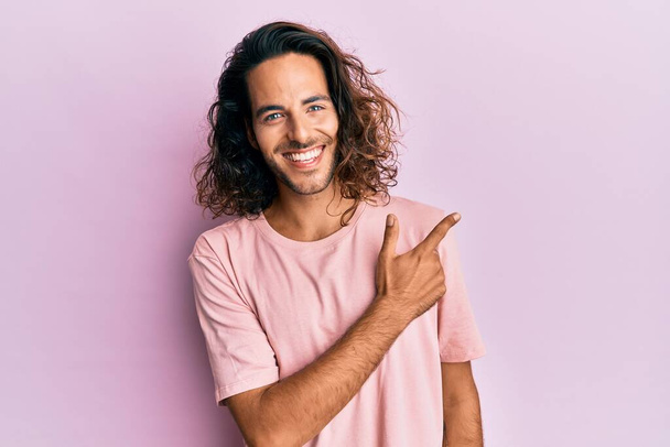 Молодой красивый мужчина с длинными волосами в повседневной одежде улыбается весело указывая рукой и пальцем в сторону  - Фото, изображение