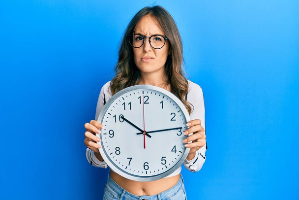 Young brunette woman holding big clock clueless and confused expression. doubt concept.  - Foto, immagini