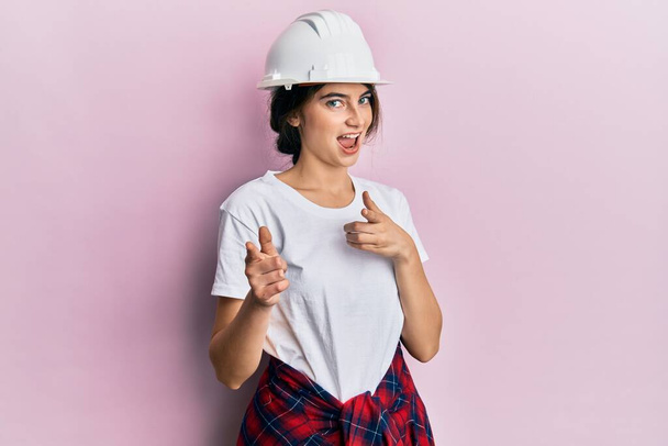 Young caucasian woman wearing hardhat pointing fingers to camera with happy and funny face. good energy and vibes.  - Фото, изображение