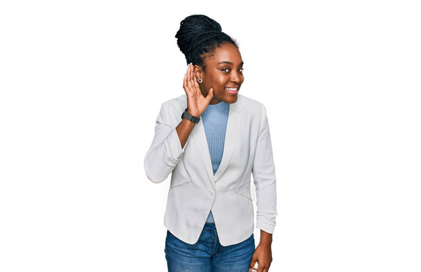 Jeune femme afro-américaine portant des vêtements de travail souriant avec la main sur l'oreille en écoutant une rumeur ou des commérages. concept de surdité.  - Photo, image