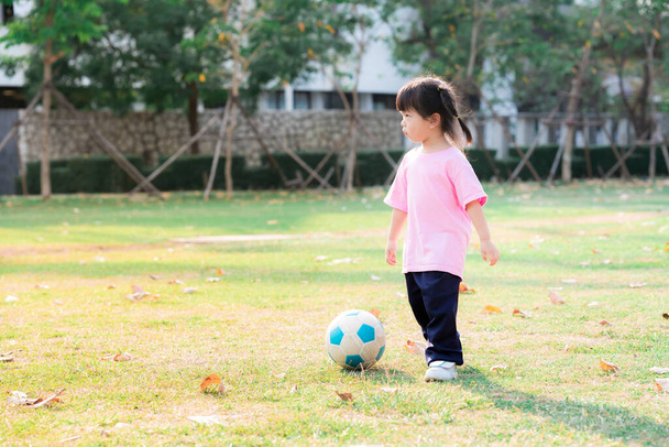 Aasialainen tyttö pelaa ja potkii palloa vihreällä nurmikolla. Söpö lapsi nauttia lämpimästä aurinkoisesta illasta. Kesällä tai keväällä. Onnelliset lapset käyttävät vaaleanpunaisia paitoja, ikä 3-4 vuotta. - Valokuva, kuva