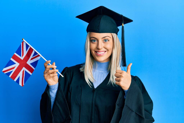 Krásná blondýnka žena na sobě maturitní čepici a slavnostní župan drží uk vlajku s úsměvem šťastný a pozitivní, palec nahoru dělá vynikající a schvalovací znamení  - Fotografie, Obrázek