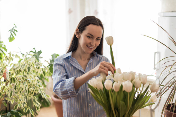 Mavi gömlekli beyaz kadın vazodaki beyaz lalelerle ilgileniyor. Doğadan esinlenilmiş ortamlar, bahar, rahat ev konsepti. - Fotoğraf, Görsel