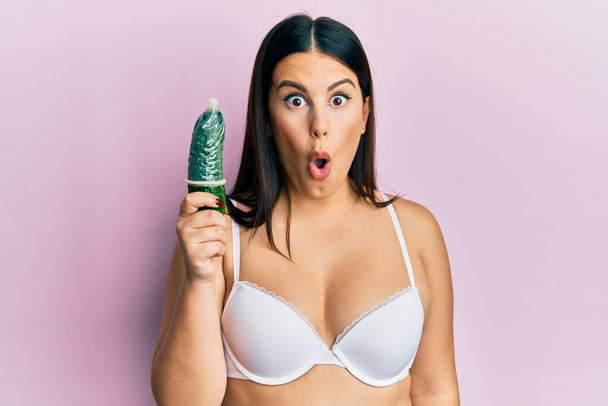 mooi brunette vrouw holding condoom op komkommer voor seks onderwijs bang en verbaasd met open mond voor verrassing, ongeloof gezicht  - Foto, afbeelding