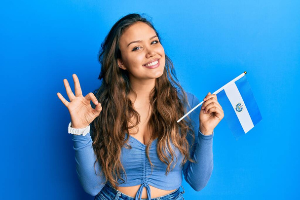 Young hispanic girl holding el salvador flag doing ok sign with fingers, smiling friendly gesturing excellent symbol  - Photo, Image