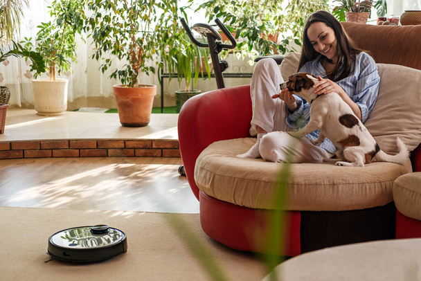 vrouw in blauw shirt speelt met hond, Jack Russell Terrier ras thuis op de bank, Robotstofzuiger op tapijt, genieten van het leven concept, gezellig en comfortabel huis - Foto, afbeelding