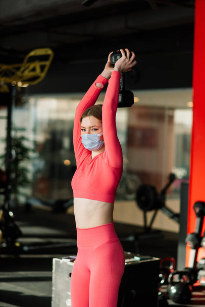 Female athlete fitness coach wearing white medical protective mask do exercises with dumbbell, invite to do fitness class in empty gym. Athletes against COVID-19 - Photo, Image