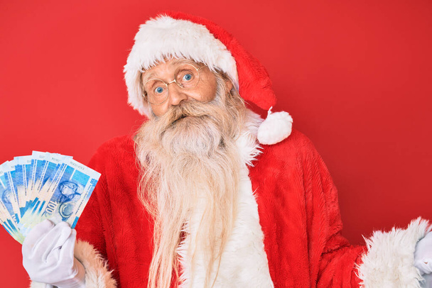 Old senior man with grey hair and long beard wearing santa claus costume holding african rands celebrating achievement with happy smile and winner expression with raised hand  - Фото, изображение