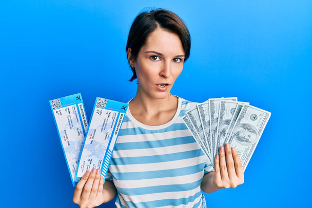 Young brunette woman with short hair holding boarding pass and dollars in shock face, looking skeptical and sarcastic, surprised with open mouth  - Photo, Image