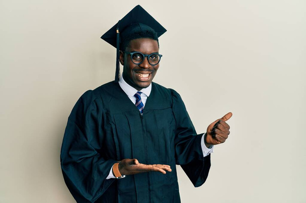 Handsome black man wearing graduation cap and ceremony robe showing palm hand and doing ok gesture with thumbs up, smiling happy and cheerful  - Foto, imagen