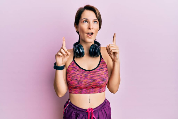 Young brunette woman with short hair wearing sportswear and headphones amazed and surprised looking up and pointing with fingers and raised arms.  - Photo, Image