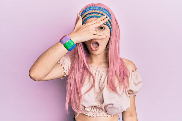 Молода іспаномовна дівчина з рожевим волоссям підглядає в шоковому покритті обличчя і очі рукою, дивлячись через пальці бояться
  - Фото, зображення