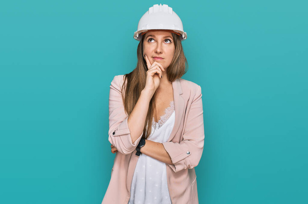 Young blonde woman wearing architect hardhat serious face thinking about question with hand on chin, thoughtful about confusing idea  - Photo, Image