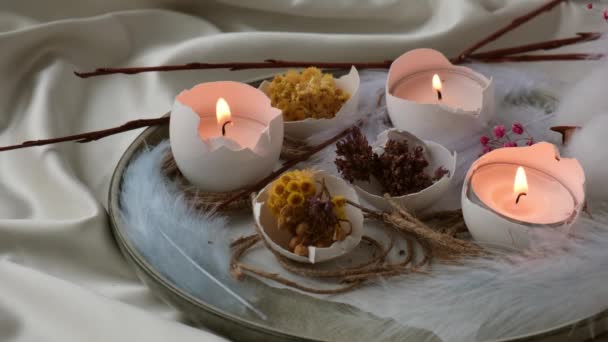 Trendige Osterdekoration. Zerbrochene Bio-Eier mit trockenen Grasblüten auf dem Teller. Kerzenlicht in der weißen Schale der Eier. Vogelfeder. Brennende Kerzen lodern Rauch auf - Filmmaterial, Video