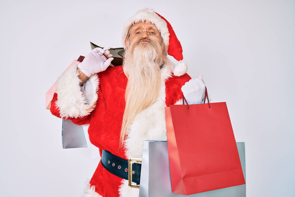 Ein alter älterer Mann mit grauen Haaren und langem Bart im Weihnachtsmann-Kostüm hält Einkaufstüten in die Kamera und bläst einen Kuss, der schön und sexy ist. Liebesausdruck.  - Foto, Bild